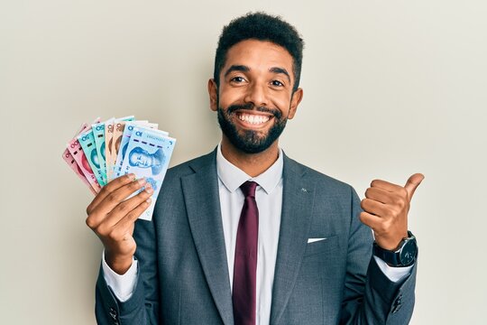Handsome hispanic man with beard wearing business suit holding yuan banknotes pointing thumb up to the side smiling happy with open mouth