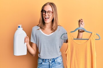 Beautiful blonde woman holding shirt on hanger and detergent bottle winking looking at the camera...