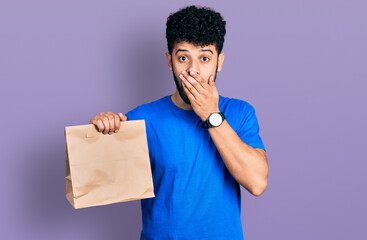 Young arab man with beard holding take away paper bag covering mouth with hand, shocked and afraid...