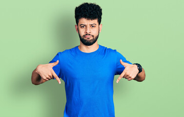 Young arab man with beard wearing casual blue t shirt pointing down looking sad and upset,...