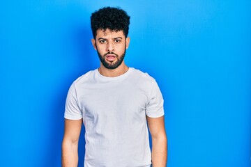 Young arab man with beard wearing casual white t shirt in shock face, looking skeptical and sarcastic, surprised with open mouth