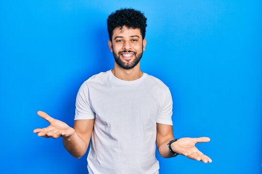 Young arab man with beard wearing casual white t shirt smiling cheerful with open arms as friendly welcome, positive and confident greetings