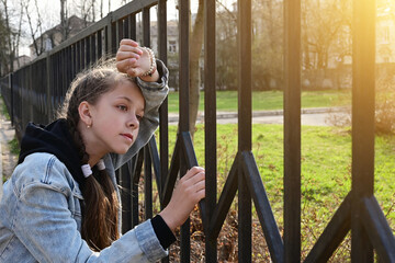 pretty, thoughtful girl in denim clothes is standing by a metal fence, leaning against it and looking into the distance. Close-up portrait in the sunlight