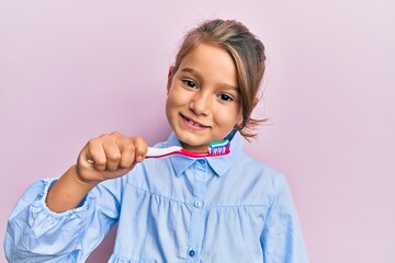 Little beautiful girl holding toothbrush with toothpaste looking positive and happy standing and smiling with a confident smile showing teeth