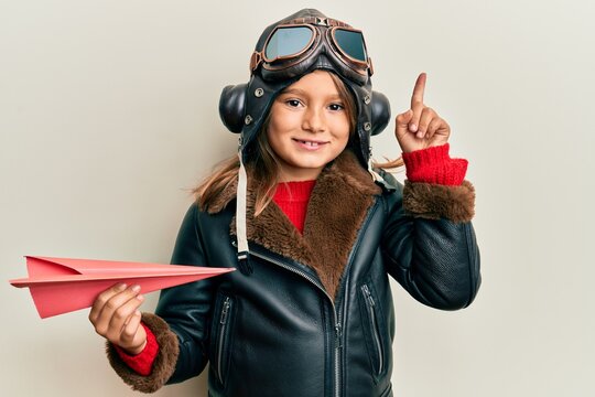 Little beautiful girl wearing pilot uniform holding paper plane smiling with an idea or question pointing finger with happy face, number one