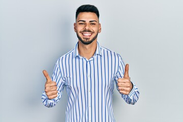 Naklejka premium Young hispanic man with beard wearing casual striped shirt success sign doing positive gesture with hand, thumbs up smiling and happy. cheerful expression and winner gesture.