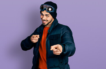 Young hispanic man with beard wearing snow wear and sky glasses pointing fingers to camera with happy and funny face. good energy and vibes.