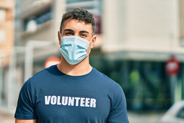 Young hispanic volunteer man wearing medical mask standing at the city.