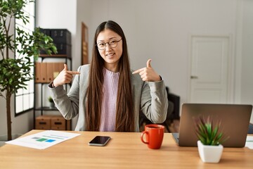 Young chinese business worker wearing business style sitting on desk at office looking confident with smile on face, pointing oneself with fingers proud and happy.