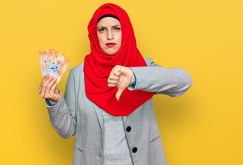 Beautiful hispanic woman wearing islamic hijab holding 10 swiss franc banknotes with angry face,...