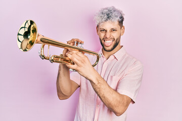 Young hispanic man with modern dyed hair playing trumpet sticking tongue out happy with funny expression. - obrazy, fototapety, plakaty