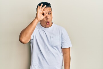 Young latin man wearing casual white t shirt doing ok gesture shocked with surprised face, eye looking through fingers. unbelieving expression.