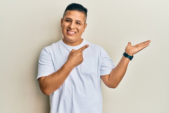 Young latin man wearing casual white t shirt amazed and smiling to the camera while presenting with hand and pointing with finger.