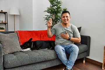 Young latin man and dog sitting on the sofa at home smiling swearing with hand on chest and fingers...