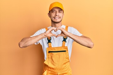 Hispanic young man wearing handyman uniform smiling in love doing heart symbol shape with hands. romantic concept.