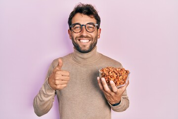 Handsome man with beard holding peanuts smiling happy and positive, thumb up doing excellent and approval sign