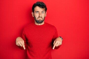 Handsome man with beard wearing casual red sweater pointing down looking sad and upset, indicating...