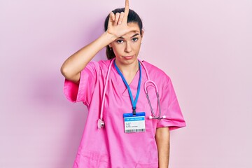 Young brunette woman wearing doctor uniform and stethoscope making fun of people with fingers on...