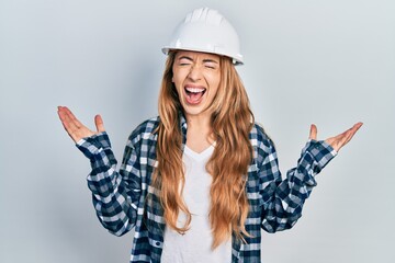 Young caucasian woman wearing architect hardhat celebrating mad and crazy for success with arms raised and closed eyes screaming excited. winner concept