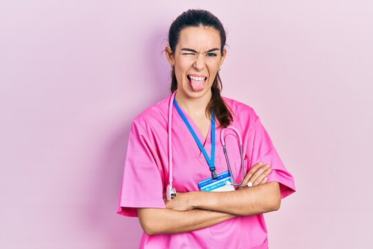 Young brunette woman wearing doctor uniform and stethoscope standing with arms crossed sticking tongue out happy with funny expression.