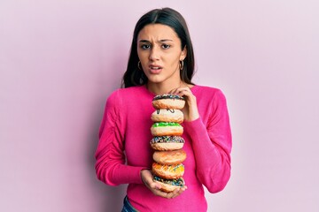 Young hispanic woman holding pile of tasty colorful doughnuts clueless and confused expression....