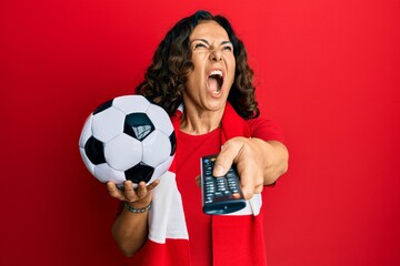 Middle age hispanic woman football hooligan holding ball using tv control angry and mad screaming...