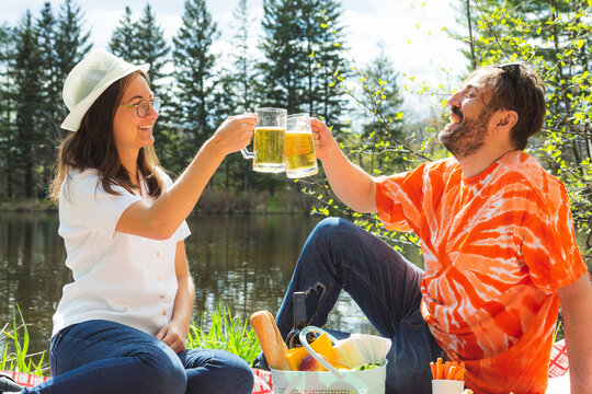 Happy young couple of friends toasting together and laughing at the park - Two friends drinking beers and having fun during pic nic barbecue - Lifestyle and people concept.