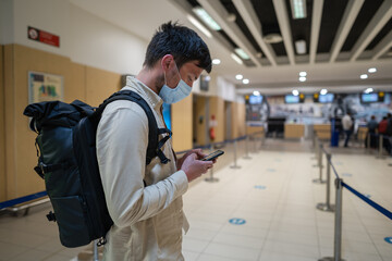Male passenger in mask with backpack uses smartphone and stands with social distance on check-in line at airport of Cyprus, paphos city. Travel by plane during quarantine coronavirus, new norm