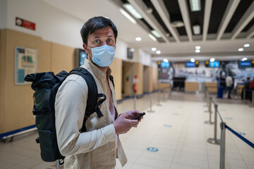 Fototapeta na wymiar Male passenger in mask with backpack uses smartphone and stands with social distance on check-in line at airport of Cyprus, paphos city. Travel by plane during quarantine coronavirus, new norm