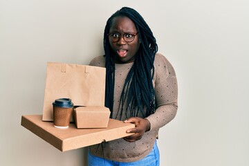 Young black woman with braids holding take away food in shock face, looking skeptical and...
