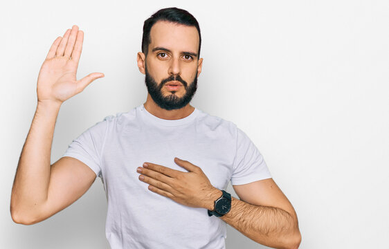 Young man with beard wearing casual white t shirt swearing with hand on chest and open palm, making a loyalty promise oath