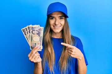 Young hispanic woman wearing delivery uniform and cap holding japanese yuan smiling happy pointing with hand and finger