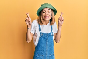 Young caucasian blonde woman wearing denim jumpsuit and hat with 90s style gesturing finger crossed...