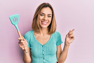 Young caucasian blonde woman holding silicone spatula smiling happy pointing with hand and finger...