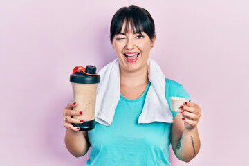 Young hispanic woman wearing sportswear holding water bottle winking looking at the camera with...