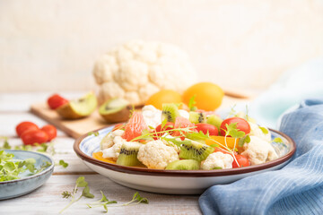 Vegetarian salad of cauliflower cabbage, kiwi, tomatoes, microgreen sprouts on gray wooden background. Side view, selective focus.