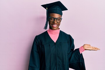 Young african american girl wearing graduation cap and ceremony robe smiling cheerful presenting and pointing with palm of hand looking at the camera.