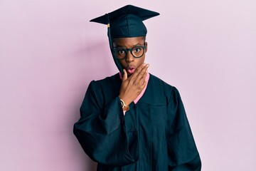 Young african american girl wearing graduation cap and ceremony robe looking fascinated with disbelief, surprise and amazed expression with hands on chin