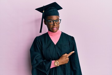 Young african american girl wearing graduation cap and ceremony robe pointing aside worried and...