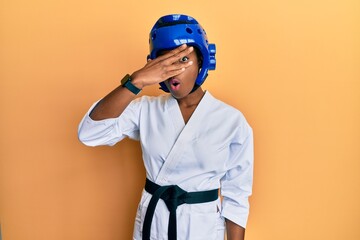 Young african american girl wearing taekwondo kimono and protection helmet peeking in shock covering face and eyes with hand, looking through fingers afraid