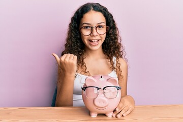 Obraz na płótnie Canvas Teenager hispanic girl holding piggy bank with glasses pointing thumb up to the side smiling happy with open mouth