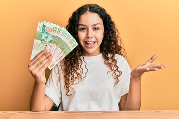 Teenager hispanic girl holding 50 hong kong dollars banknotes celebrating achievement with happy smile and winner expression with raised hand