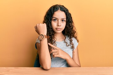 Teenager hispanic girl wearing casual clothes sitting on the table in hurry pointing to watch time,...