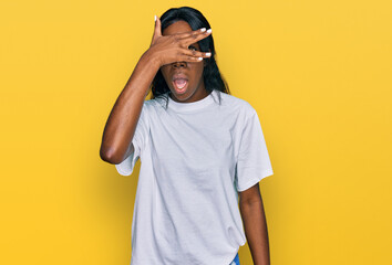 Fototapeta premium Young african american woman wearing casual white t shirt peeking in shock covering face and eyes with hand, looking through fingers with embarrassed expression.