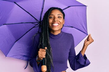 African american woman holding purple umbrella screaming proud, celebrating victory and success...