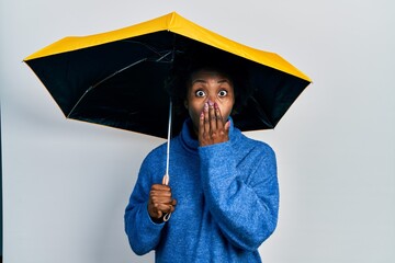 Young african american woman holding yellow umbrella covering mouth with hand, shocked and afraid...