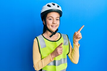 Beautiful brunette little girl wearing bike helmet and reflective vest smiling and looking at the camera pointing with two hands and fingers to the side.