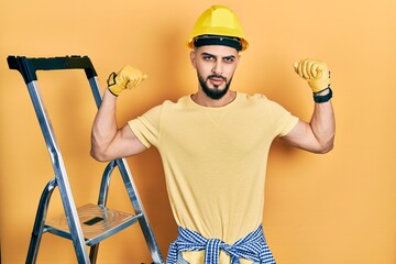 Handsome man with beard by construction stairs wearing hardhat showing arms muscles smiling proud. fitness concept.