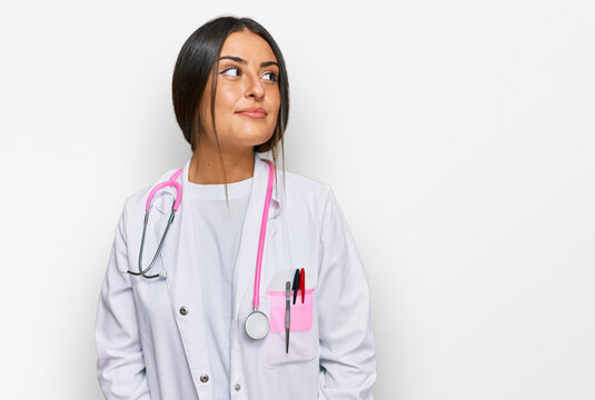 Beautiful hispanic woman wearing doctor uniform and stethoscope smiling looking to the side and staring away thinking.