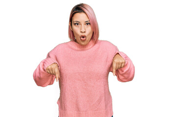 Hispanic woman with pink hair wearing casual winter sweater pointing down with fingers showing advertisement, surprised face and open mouth